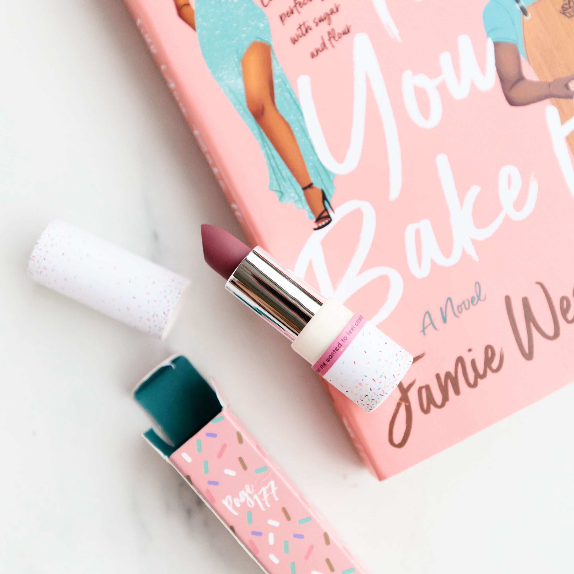 A copy of Fake It Till You Bake It by Jamie Wesley is in the corner. A tube of dark pink lipstick sits opened on top of the book cover. The tube is white and covered in rainbow dots. A pink box with a pattern of multi-colored sprinkles sits opened beneath the lipstick with Page 177 written on it.