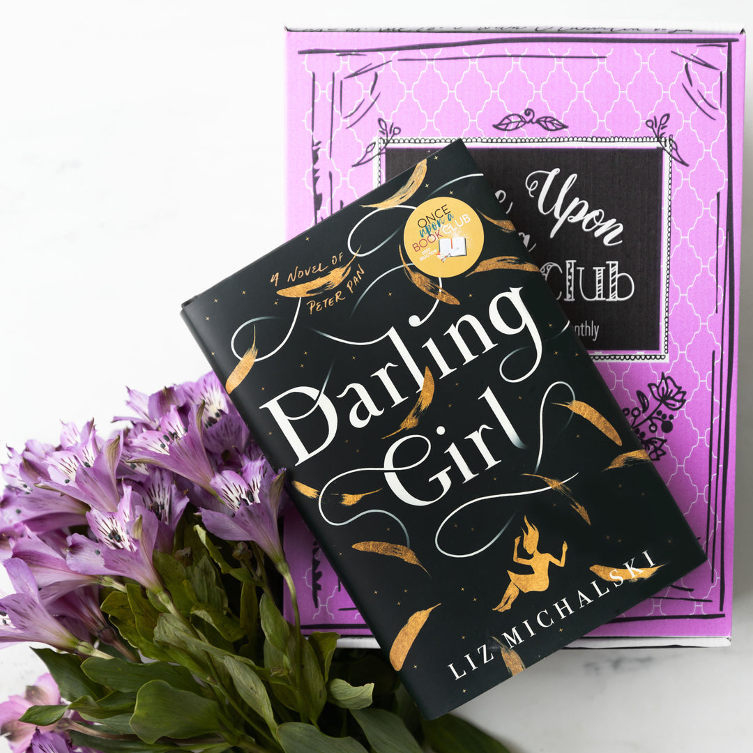 a hardcover edition of Darling Girl lays on a pink box. Underneath the book and box are purple flowers