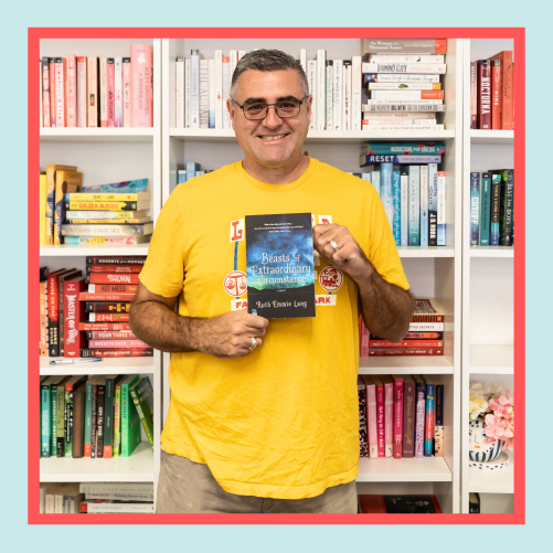 a white man wearing glasses stands in front of a bookshelf wearing a yellow shirt, smiling and holding a book