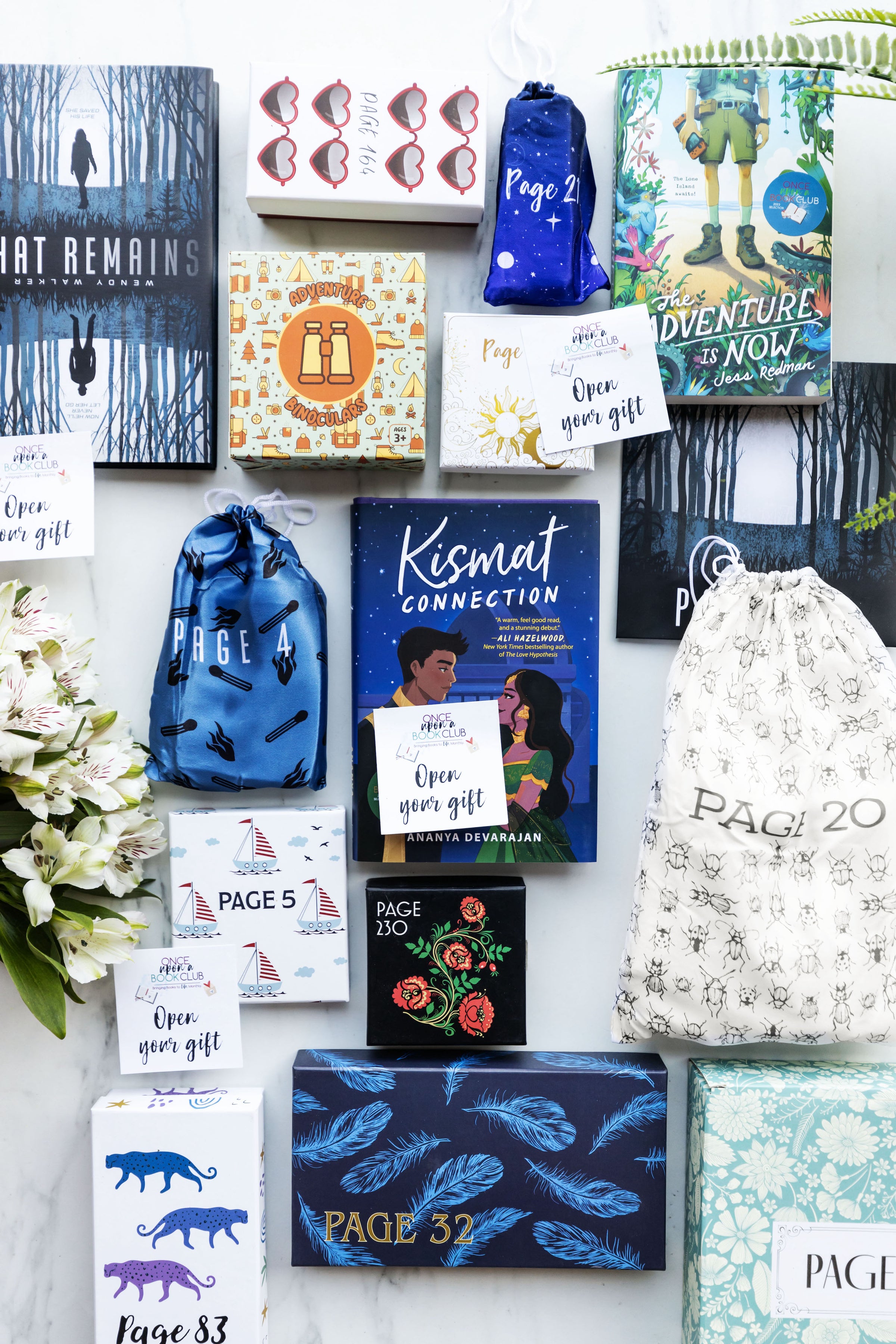A collection of books and wrapped gifts in a flatlay style. Books include What Remains by Wendy Walker (the exclusive Once Upon a Book Club edition), The Adventure is Now by Jess Redman, and The Kismat Connection by Ananya Devarajan. Flowers are on the left side of the image. Gift bags are all labeled with various page numbers. There are also Open Your Gift sticky notes scattered around the image.