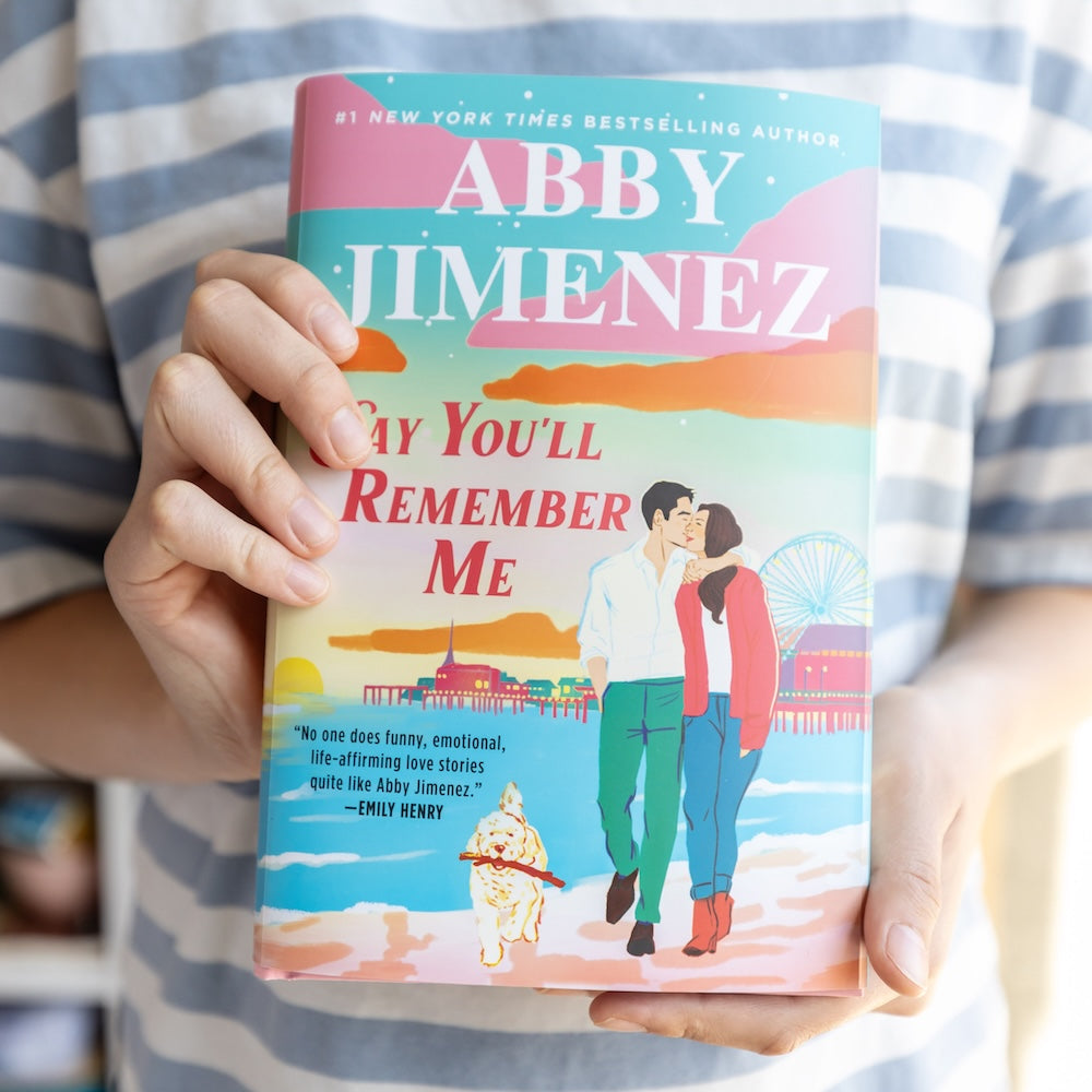 'Say You'll Remember Me' by Abby Jimenez held by a person showing side 1 of the dust jacket featuring the standard artwork.