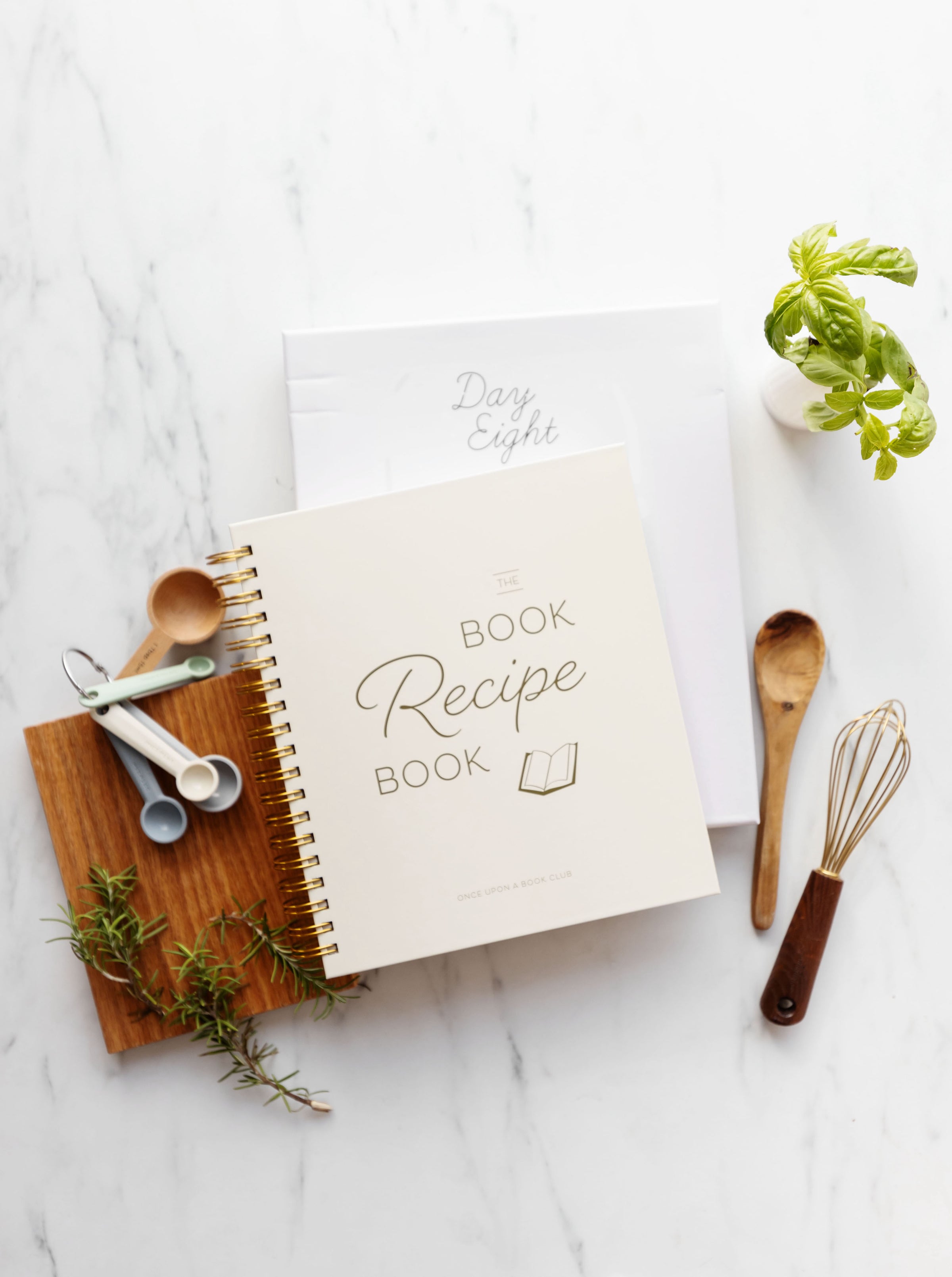 A wooden-covered Book Recipe Book sits on a white marble counter. It is surrounded by greenery, a wooden cutting board, a set of measuring spoons, a whisk, and a wooden spoon.