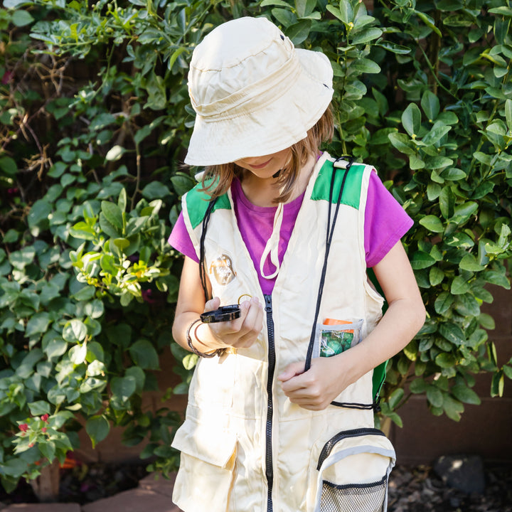 a girl wears a tan safari hat and vest against a background of green plants, holding a magnifying glass and looking down