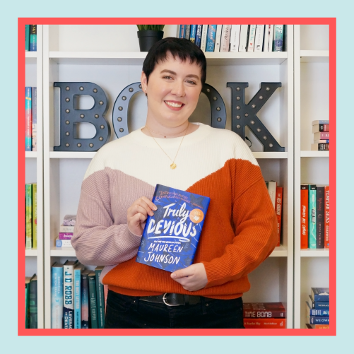 a white woman with short black hair wearing a sweater stands in front of a bookshelf, smiling and holding a book