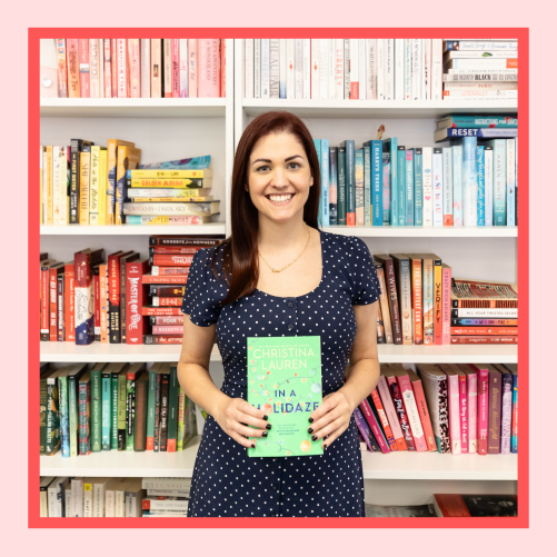 a white woman with dark red hair wearing a blue dress with white polka-dots stands in front of a bookshelf, smiling and holding a book