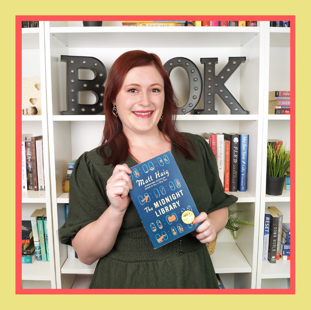 a white woman wearing a green dress with red hair stands in front of a bookshelf, smiling and holding a book