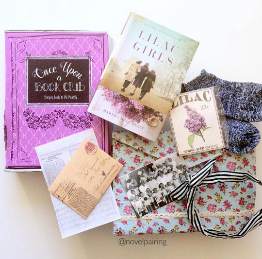 a hardcover edition of Lilac Girls is on a pink box. Surrounding the box and book are a variety of gifts including lilac seeds, socks, an envelope, and a black and white photo