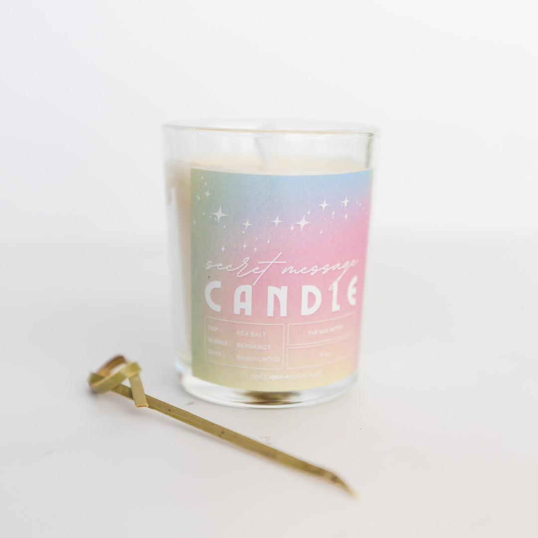 A small candle with a wooden pick next to it. The candle label is in pastel colors and says Secret Message Candle.