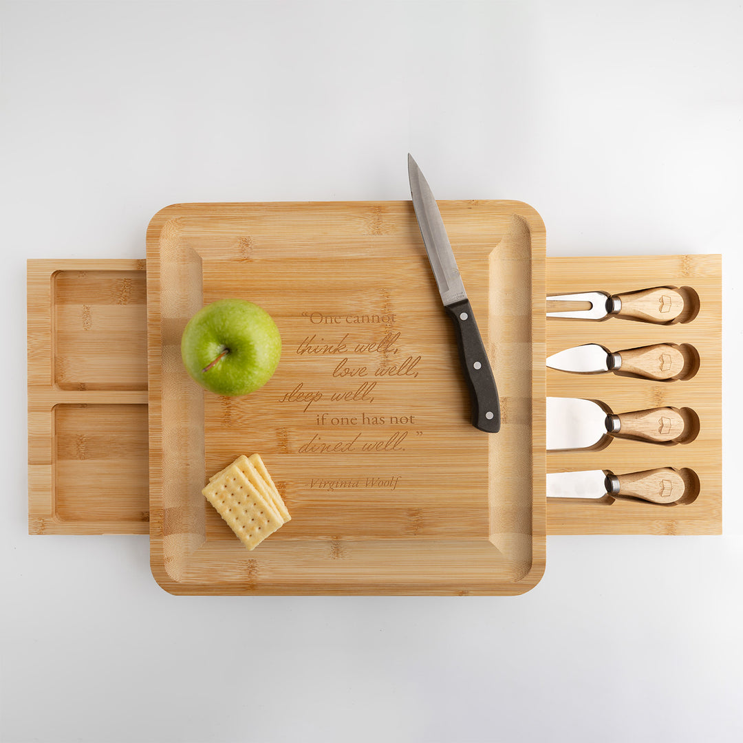 A wooden charcuterie board with drawers on the left and right, and a knife, apple, and crackers on it. The right drawer contains four cheese knives with a book icon on each wooden handle and the left drawer has two empty compartments. There is a quote on the board that says 'One cannot think well, live well, sleep well, if one has not dined well.' Virginia Woolf.