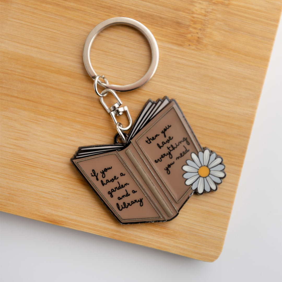 An open brown book keychain with a sunflower on it. The book 'covers' have the quote: if you have a garden and a library then you have everything you need.