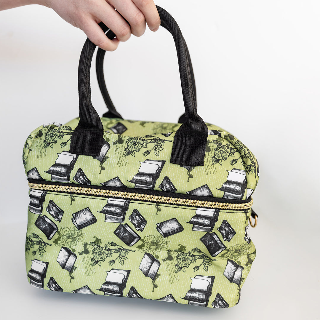 A hand holds a light green lunch box with black handles. There is a pattern of books and flowers across the green.