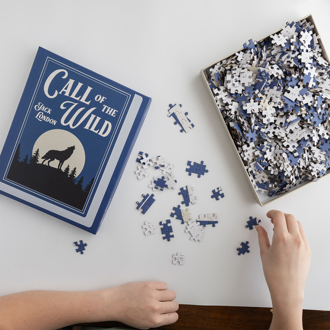 A person holds a few puzzle pieces in front of an open box filled with puzzle pieces. The top of the box is to the side and says Call of the Wild Jack London with an image of a wolf in front of a full moon.