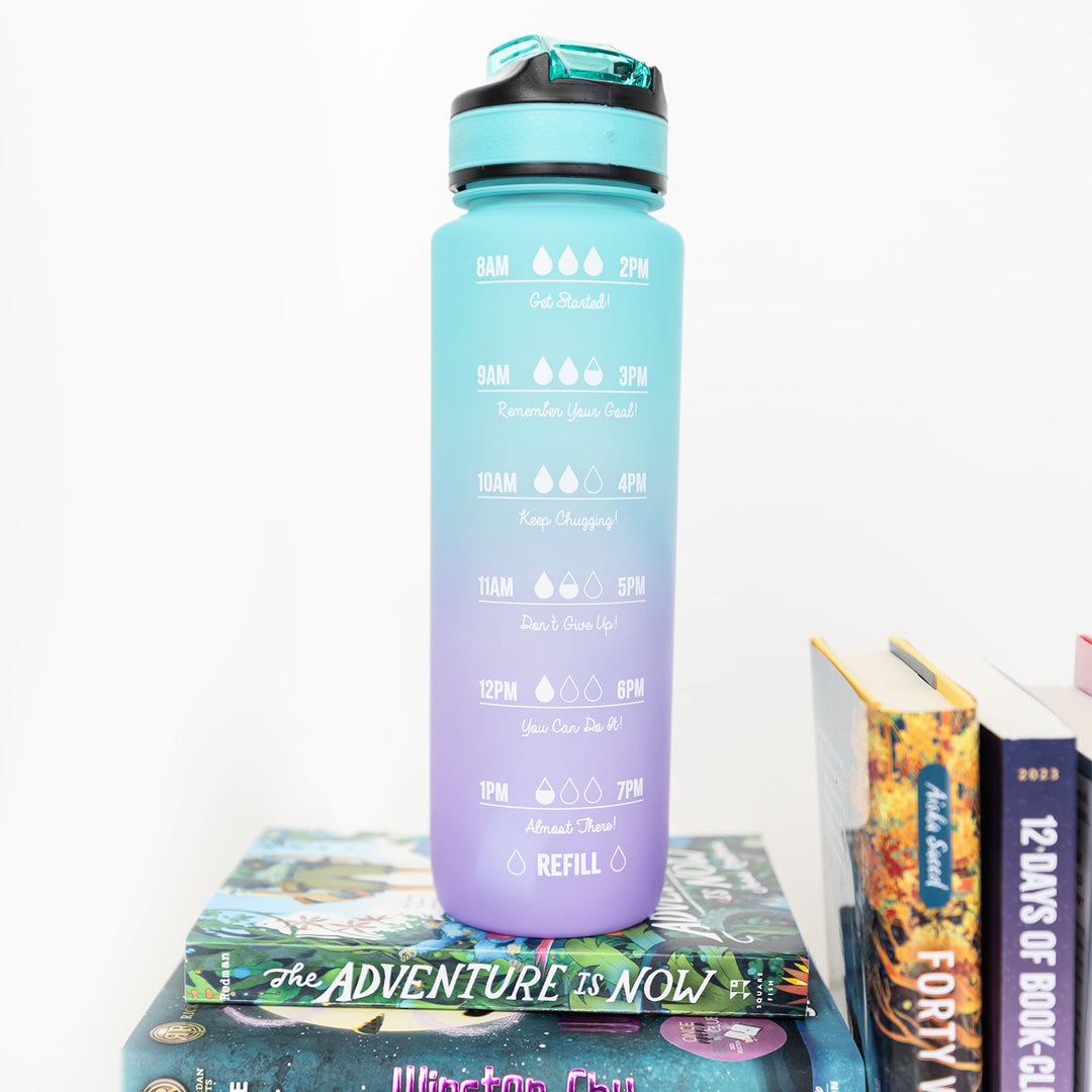A blue and purple ombre water bottle with goals to drink more water printed on the side sits atop a stack of books.
