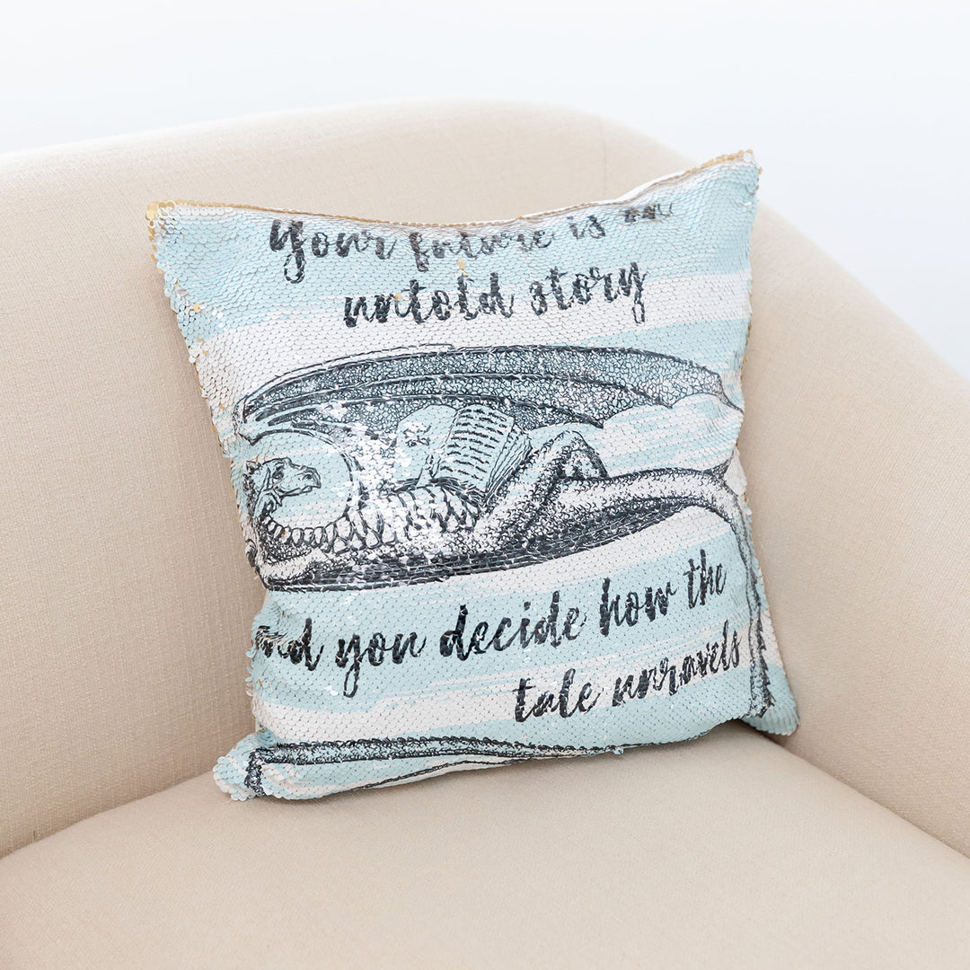 A blue and white square sequin pillow with a dragon holding a book and the quote "Your future is an untold story and you decide how the tale unravels" in a pink chair.