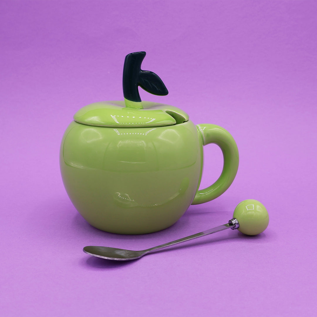 A green mug in the shape of an apple, with a lid with a black stem on top. Next to the mug is a silver spoon with a green ball at the end of the handle.