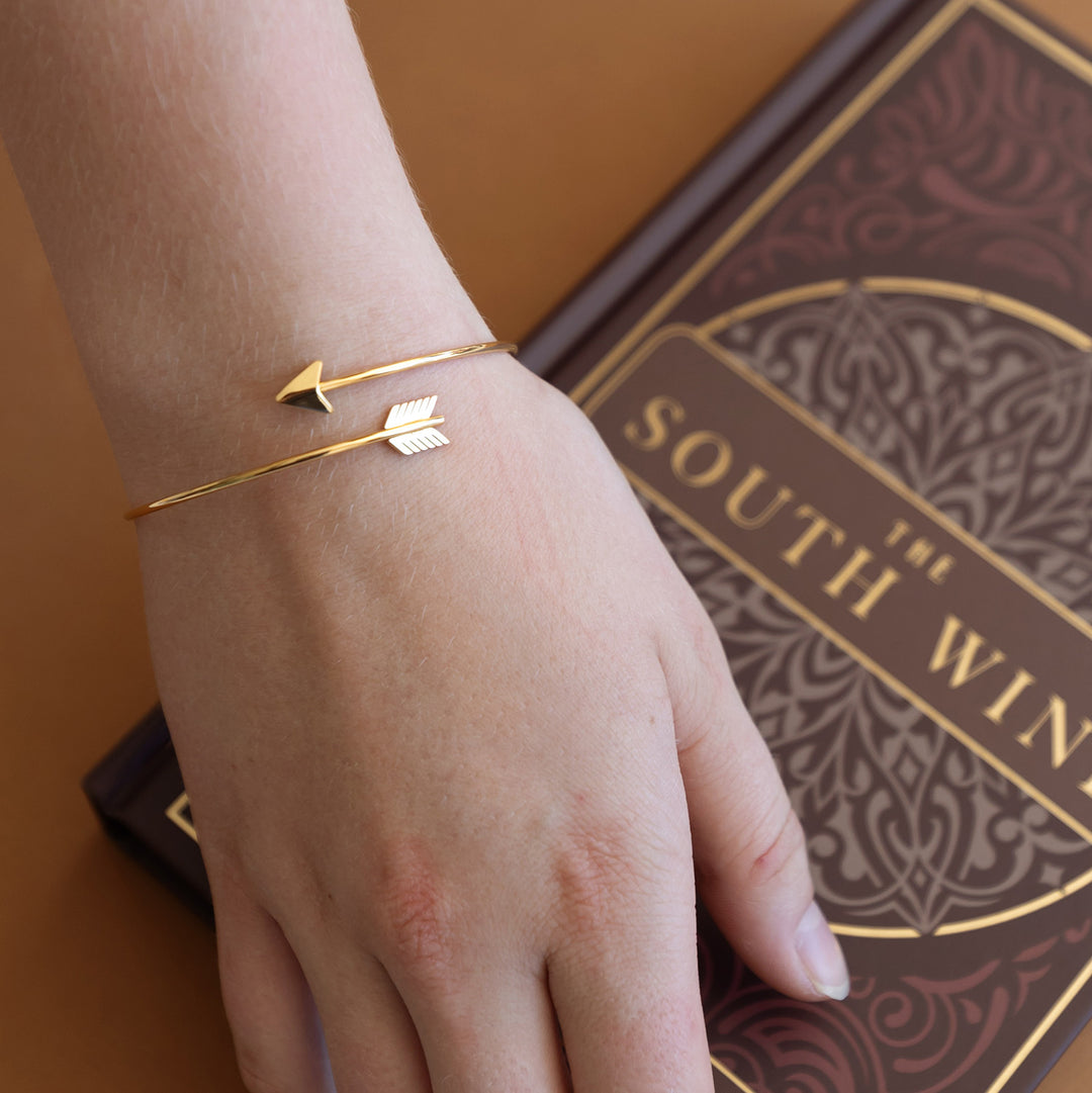 A hand lays on a special edition of The South Wind, wearing a gold arrow bracelet on her wrist.