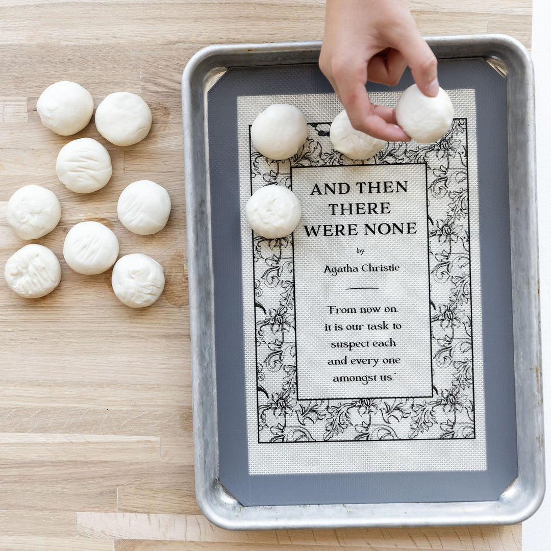a baking mat that says "And Then There Were None" by Agatha Christie on a wooden table next to a bunch of pastry balls. A white hand reaches over the baking mat