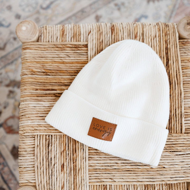 a white beanie with a brown square at the front that says "Love is energy" on a wicker table