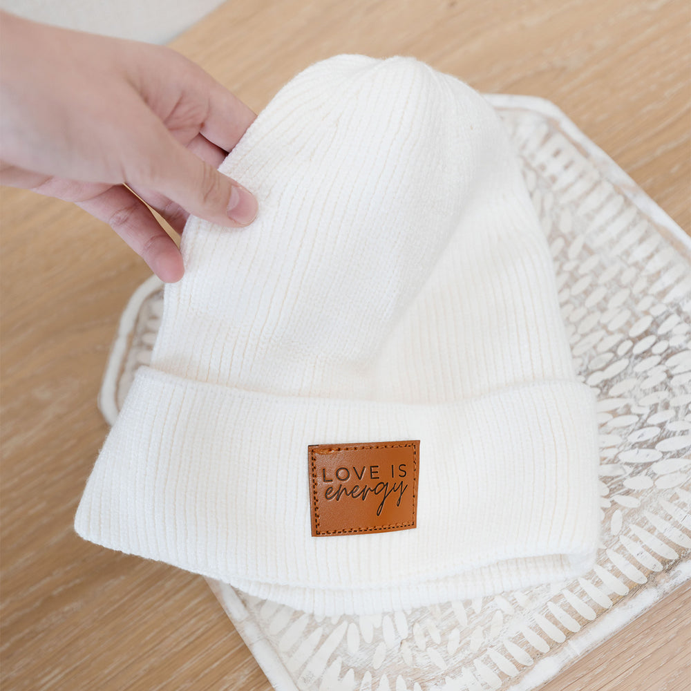 a white hand holds a white beanie with a brown square at the front that says "Love is energy"