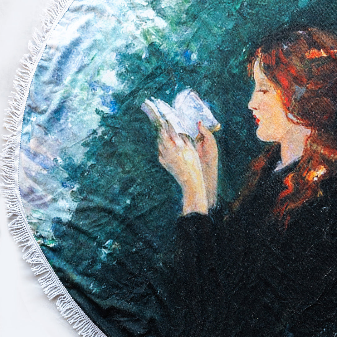 A stunning round beach blanket showing a red-headed woman reading a book on against a green and blue background. Also includes white tassels.