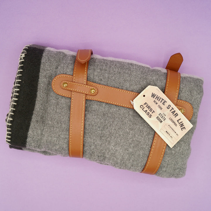 The grey plaid blanket sits wrapped in faux leather straps on a purple background.