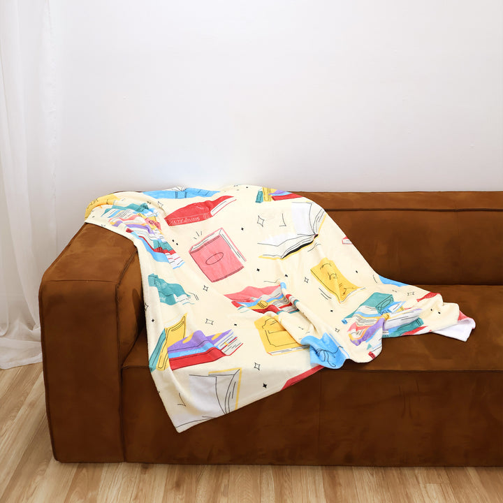 Colorful butter yellow blanket with a book design draped over a brown couch against a white wall.