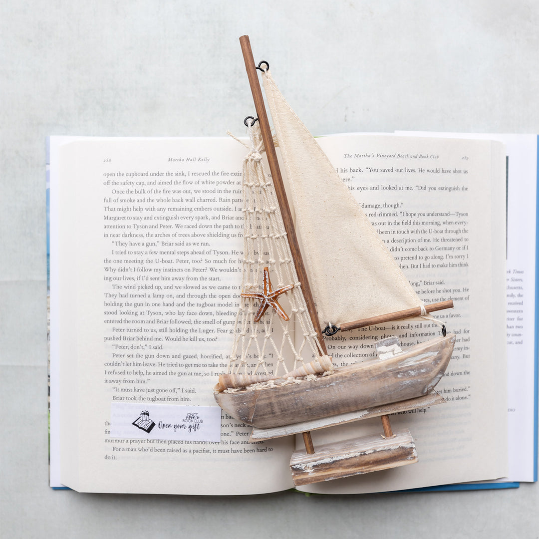 A wooden sailboat replica laying on an open book with an Open Your Gift sticky note on the page.