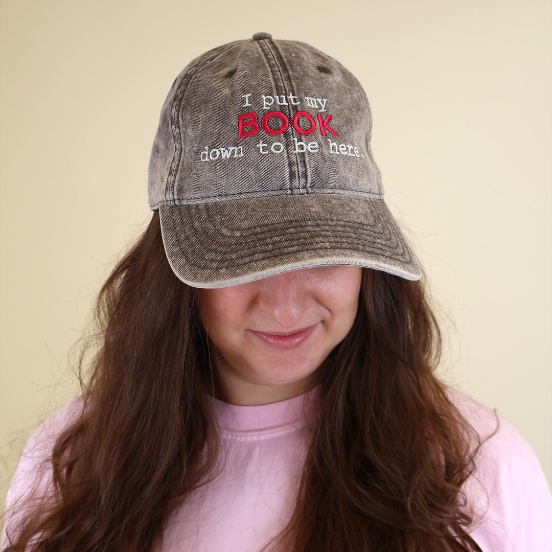 A woman wearing a grey cap with embroidered text 'I put my BOOK down to be here' on a plain background