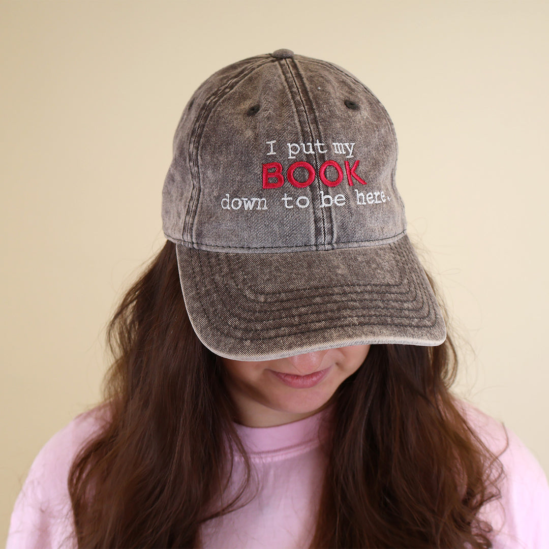 A woman wearing a grey cap with embroidered text 'I put my BOOK down to be here' on a plain background
