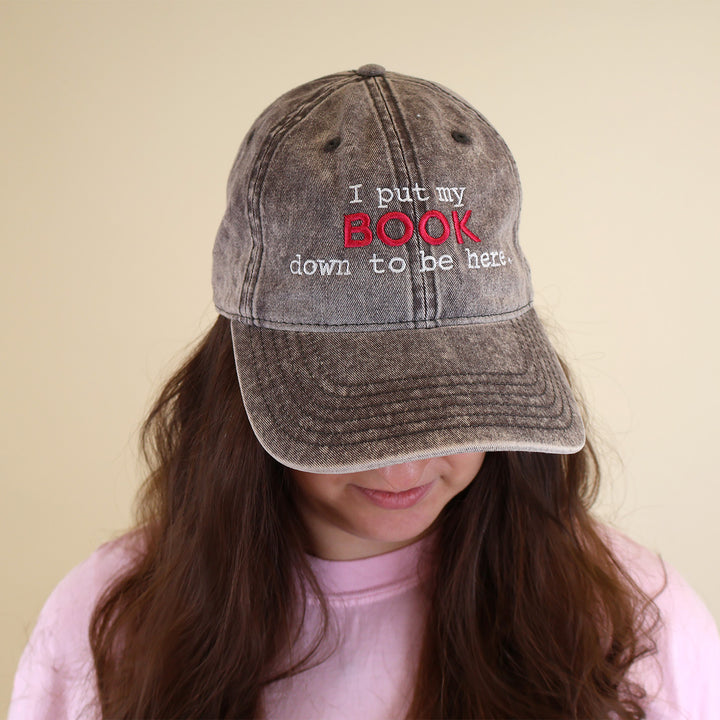 A woman wearing a grey cap with embroidered text 'I put my BOOK down to be here' on a plain background
