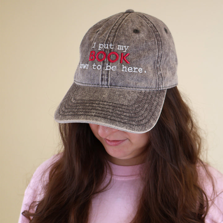 A woman wearing a grey cap with embroidered text 'I put my BOOK down to be here' on a plain background
