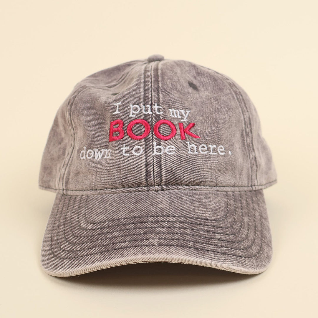 a grey cap with embroidered text 'I put my BOOK down to be here' on a plain background