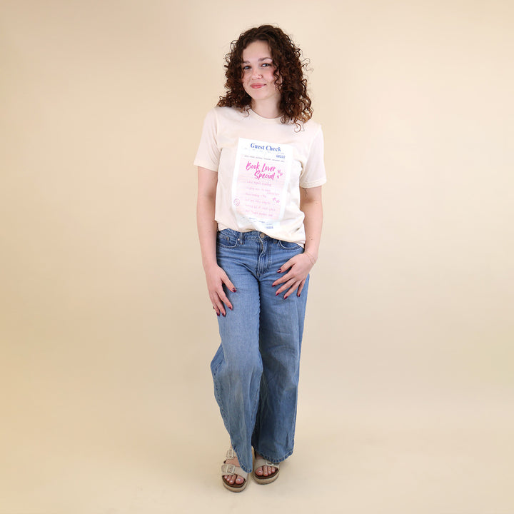 Woman wearing a cream t-shirt with a printed 'Book Lover Special' Guest Check design and jeans on a beige background