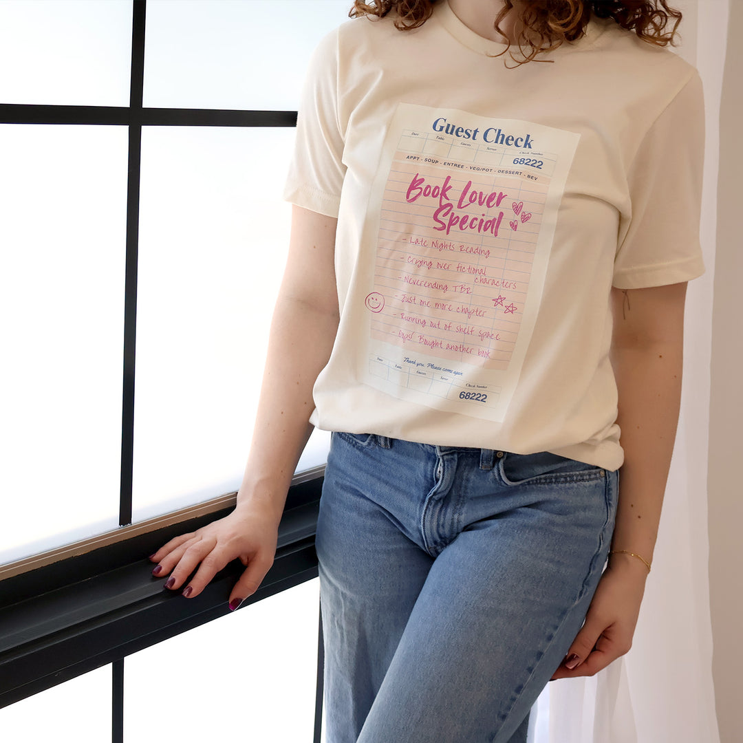 Woman wearing a cream t-shirt with a printed 'Book Lover Special' Guest Check design standing by a window
