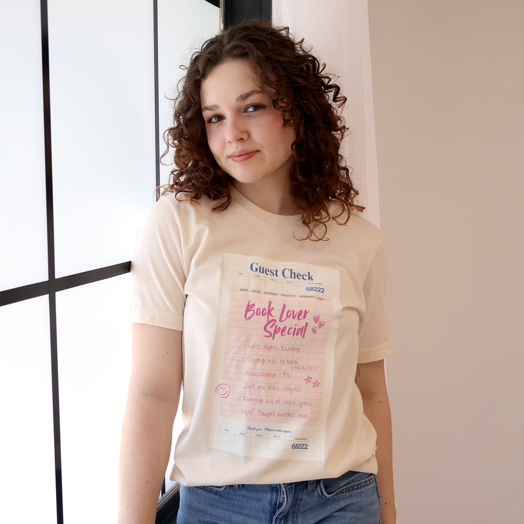 Woman wearing a cream t-shirt with a printed 'Book Lover Special' Guest Check design on a beige background