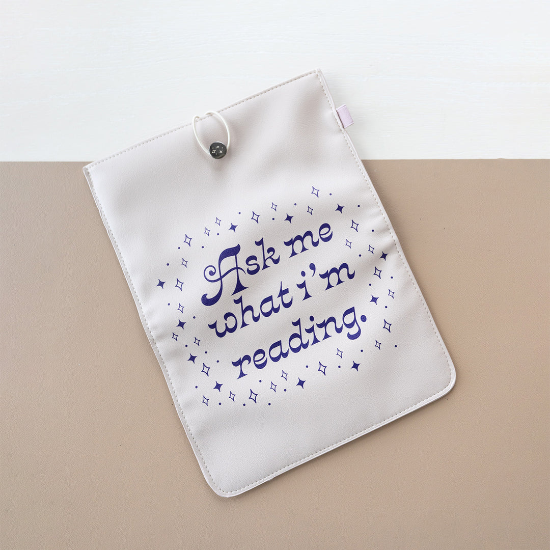 A booksleeve sits on a two-toned background. This booksleeve reads "Ask me what I'm reading" in purple font on a cream background surrounded by purple sparkles.