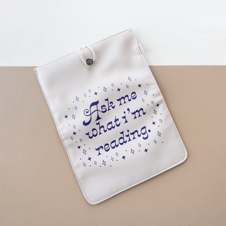 A booksleeve sits on a two-toned background. This booksleeve reads "Ask me what I'm reading" in purple font on a cream background surrounded by purple sparkles.
