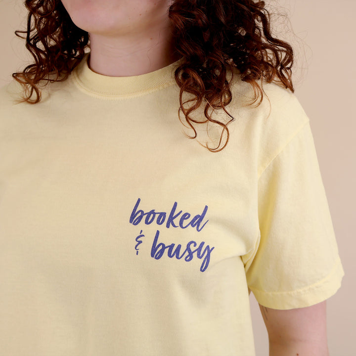 A woman wearing yellow t-shirts with 'Booked & Busy' on the front and 'Ask Me What I'm Reading' on the back surrounded by book-themed doodles.
