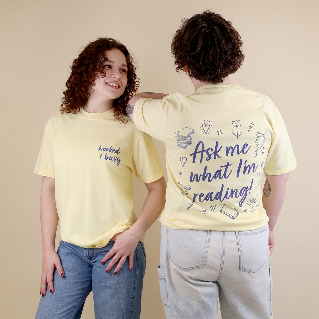 Two women wearing butter yellow t-shirts with 'Booked & Busy' on the front and 'Ask Me What I'm Reading' on the back surrounded by book-themed doodles.