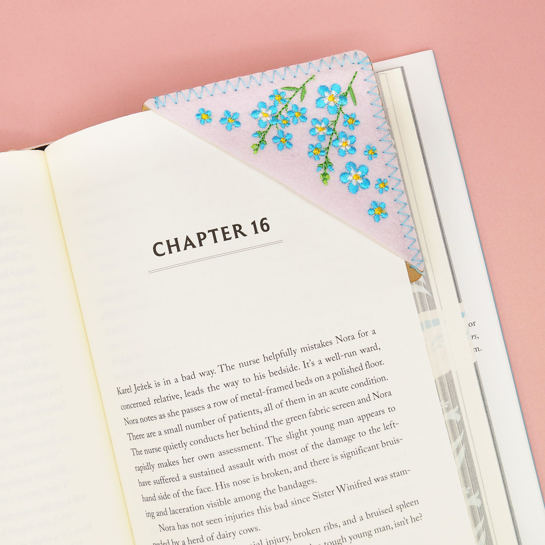 An open book with a corner bookmark on it. The corner bookmark is light pink with blue and yellow flowers on it.