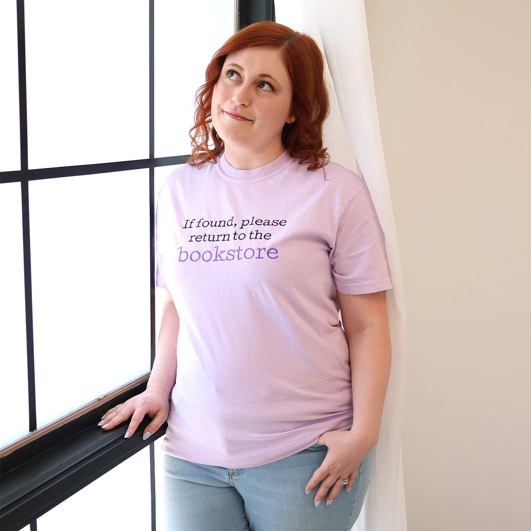 Woman wearing a light purple t-shirt with 'If found, please return to the bookstore' embroidered on it, standing by a window.