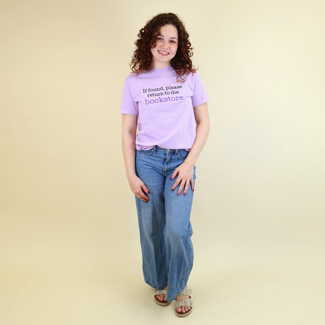 A woman wearing a light purple t-shirt with 'If found, please return to the bookstore' embroidered on it, standing on a beige background.