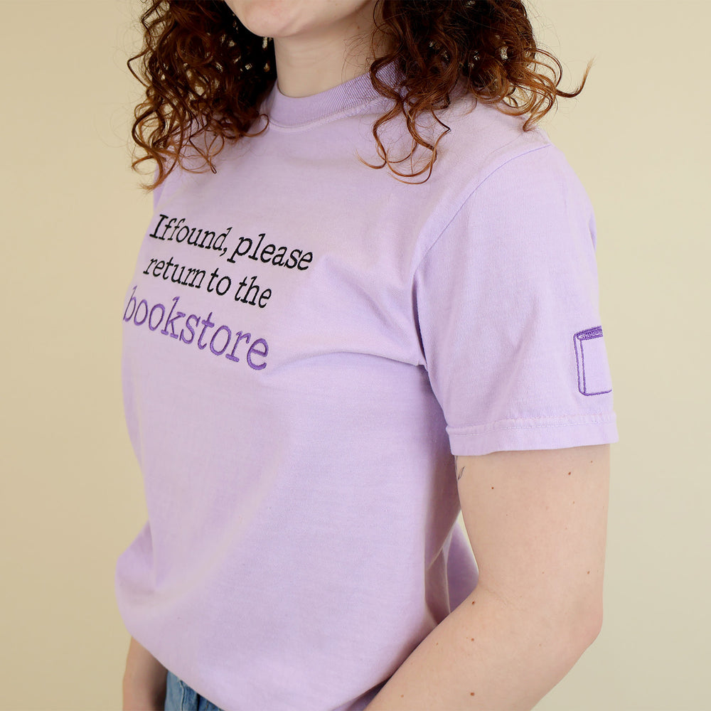Woman wearing a light purple t-shirt with 'If found, please return to the bookstore' embroidered on it and a book embroidered on the sleeve standing on a beige background.