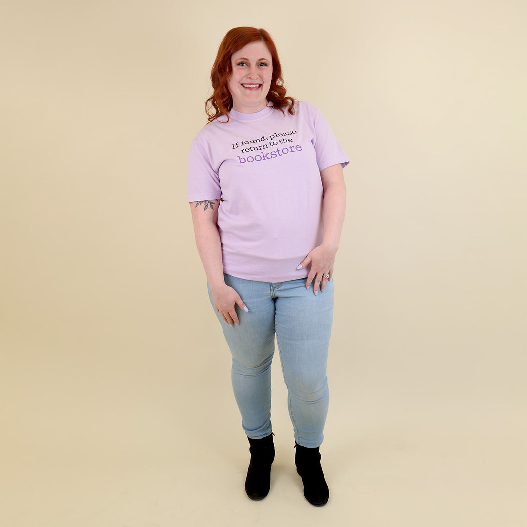 A woman wearing a light purple t-shirt with 'If found, please return to the bookstore' embroidered on it, standing on a beige background.
