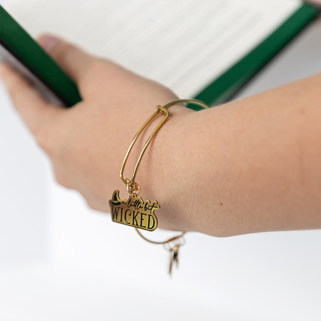 A white hand holds an open book, wearing a gold bangle bracelet with a charm that says 'a little bit WICKED' next to a witch's hat icon.
