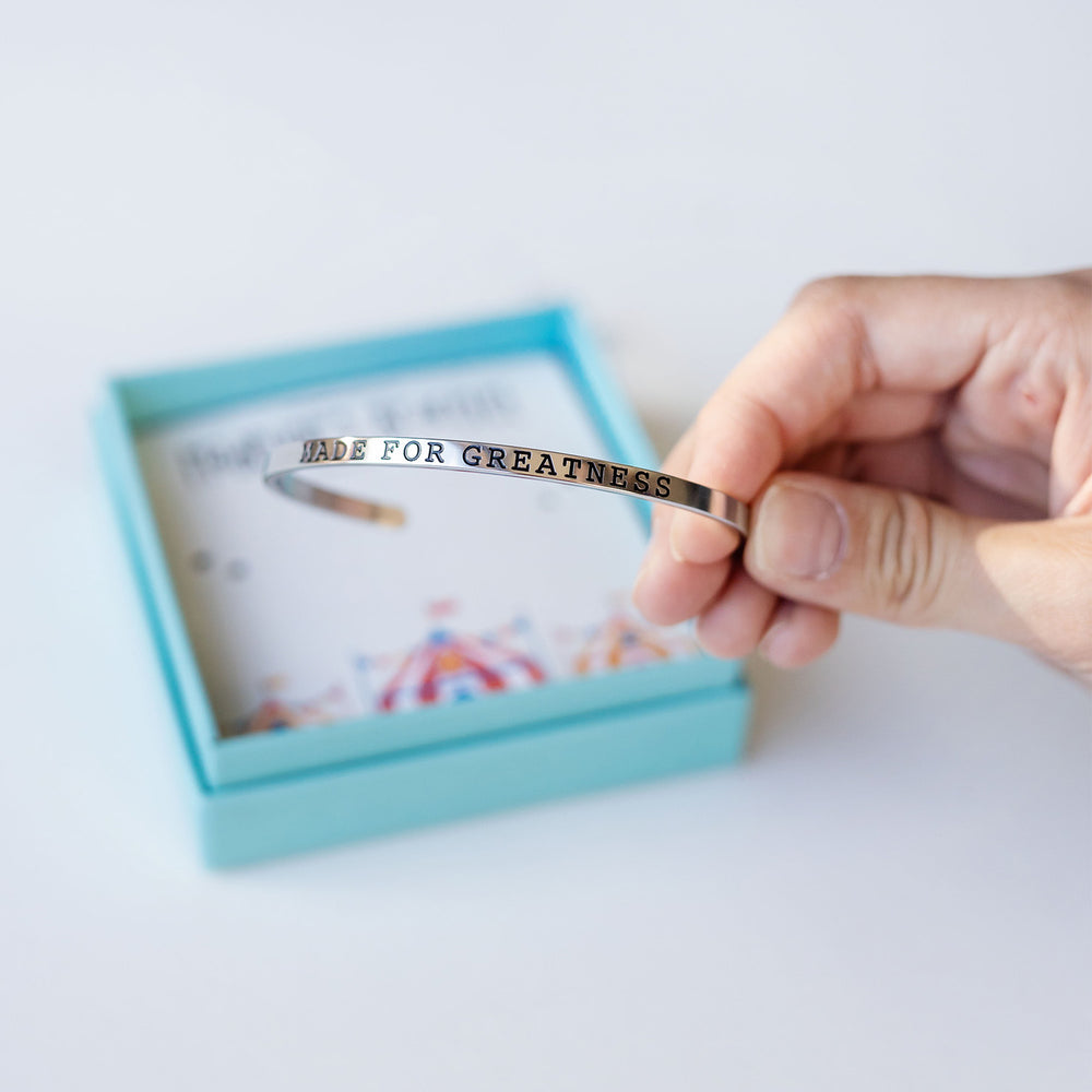 a silver cuff bracelet embossed with the words "Made for Greatness" in black lettering is held up to the camera with its box in the background.