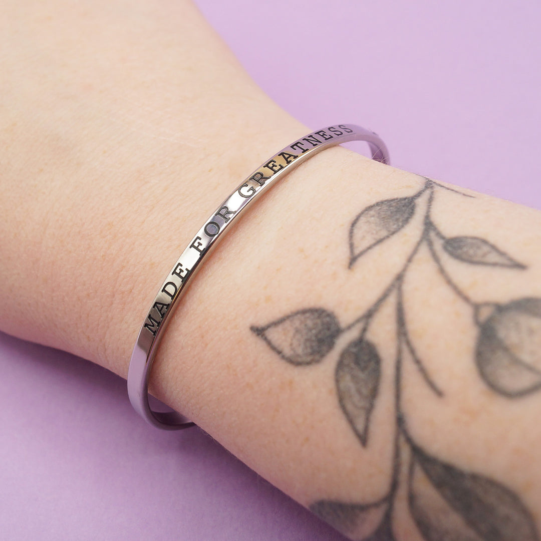 a silver cuff bracelet embossed with the words "Made for Greatness" is worn on someone's wrist.
