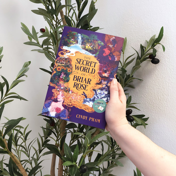 A hand holds the book 'The Secret World of the Briar Rose' by Cindy Pham, featuring a vibrant purple cover with an intricate fantasy illustration, in front of a potted olive tree against a white wall.
