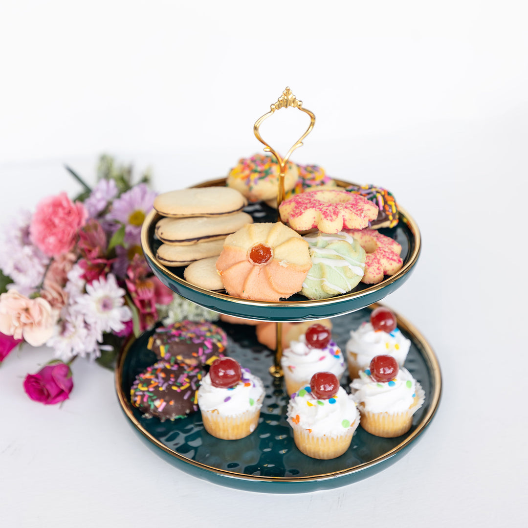 A two-tier dark green cake stand with gold accents filled with cupcakes and cookies on each level. A bouquet of flowers is laying behind the cake stand.