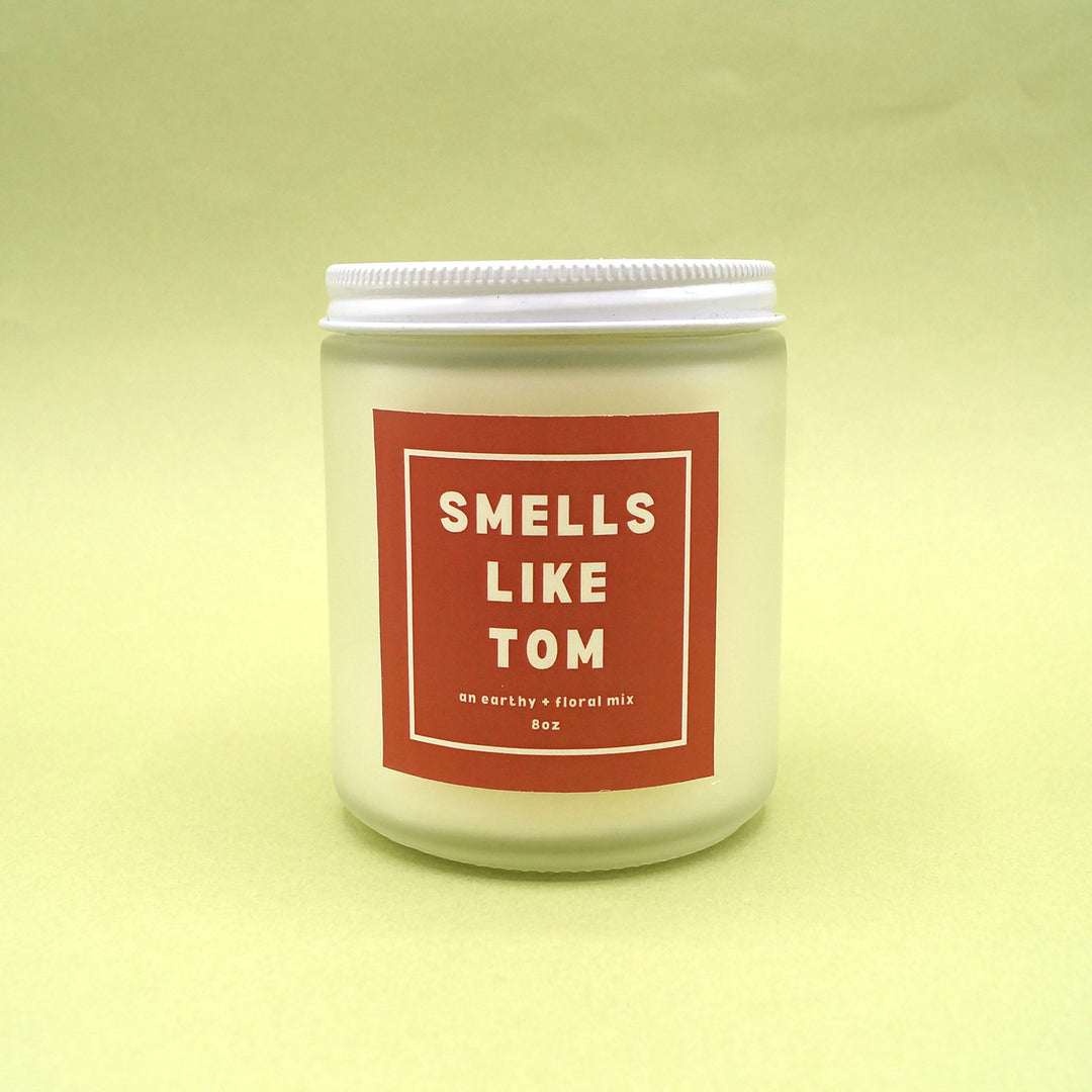 An 8oz glass candle with metal lid. The label reads "Smells Like Tom" and is inspired by Tom from the book, The Getaway List by Emma Lord. An earthy and floral scent.